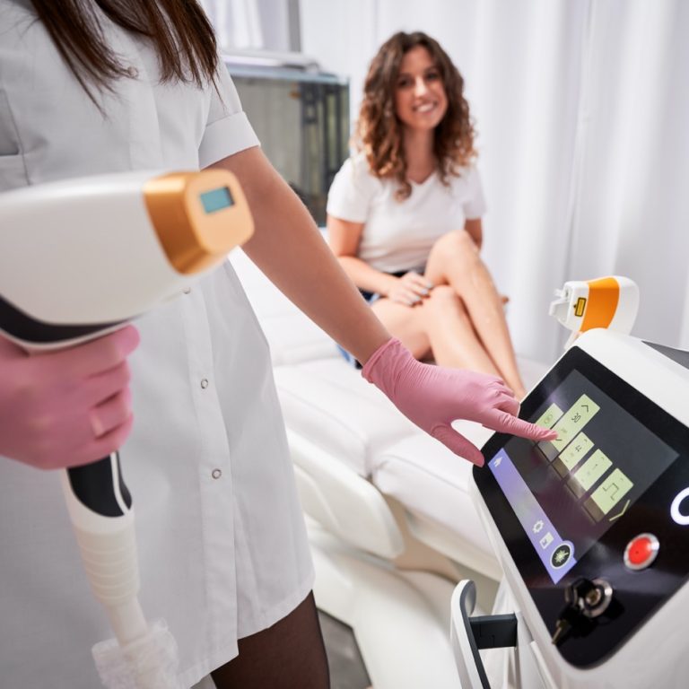 Close up of female beautician in sterile gloves using diode laser hair removal machine while smiling woman sitting on daybed. Esthetician preparing equipment for epilation procedure.